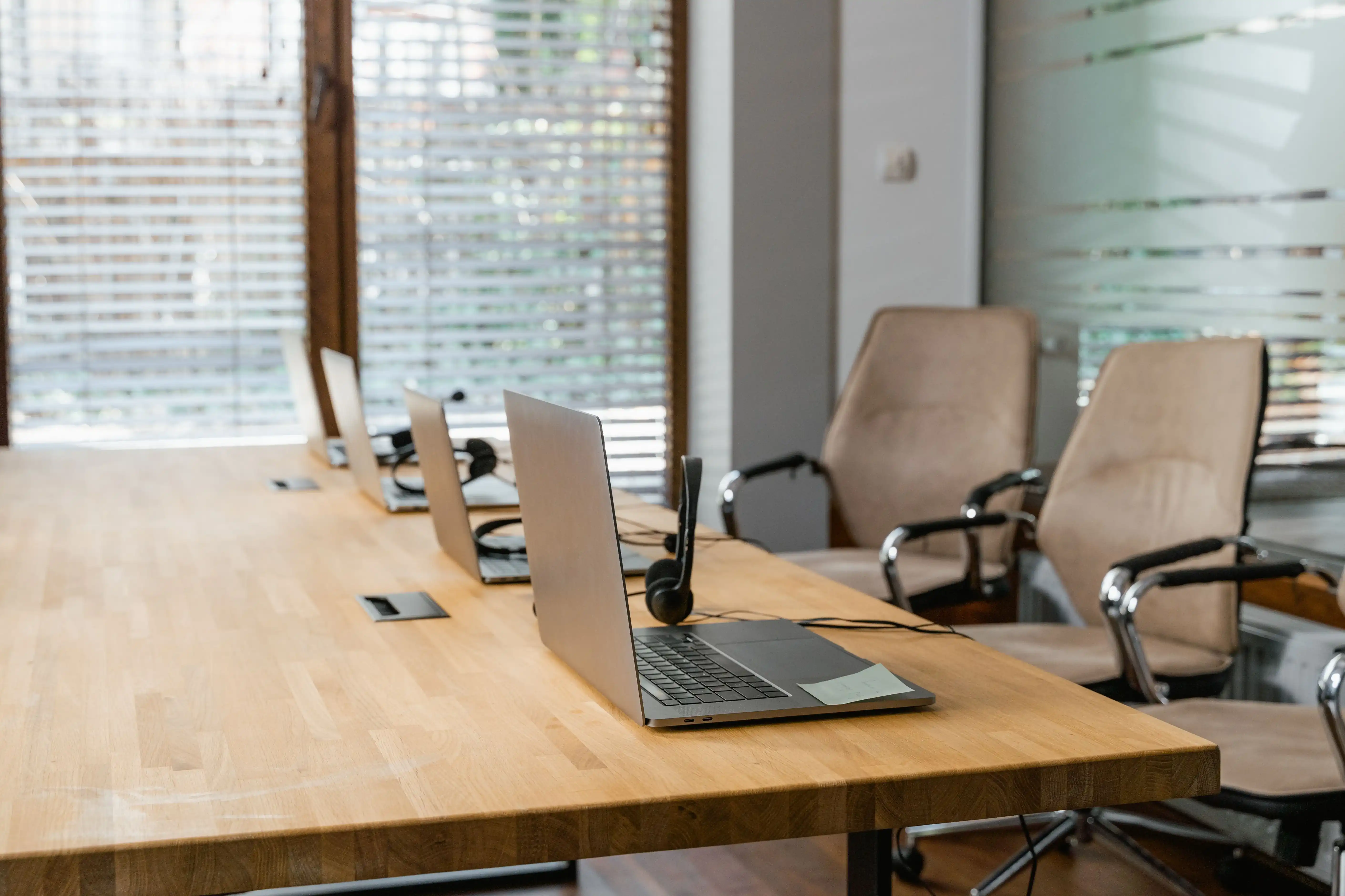 Helles Büro mit Laptops und Headsets auf einem Holztisch sowie beigen Bürostühlen